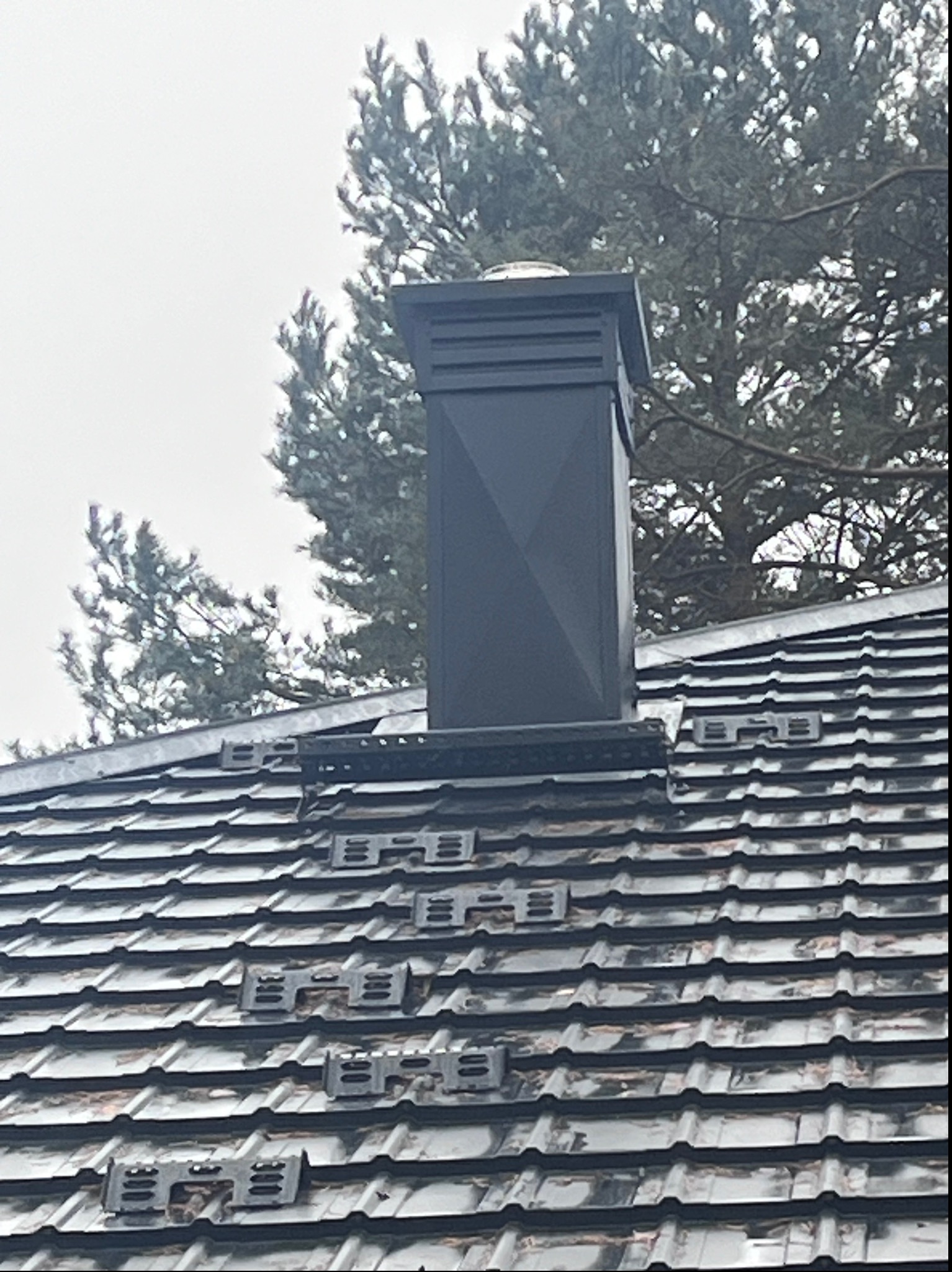 Czarny, nowoczesny komin na dachu pokrytym ciemną dachówką ceramiczną z zamkami, w tle drzewa iglaste i pochmurne niebo.