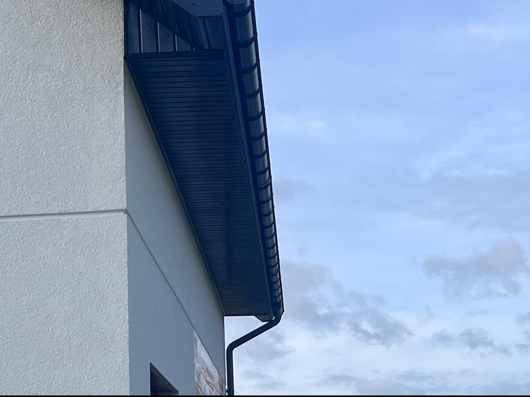 Fragment budynku z zamontowaną, ciemną podbitką dachową i rynną, na tle jasnego, pochmurnego nieba.