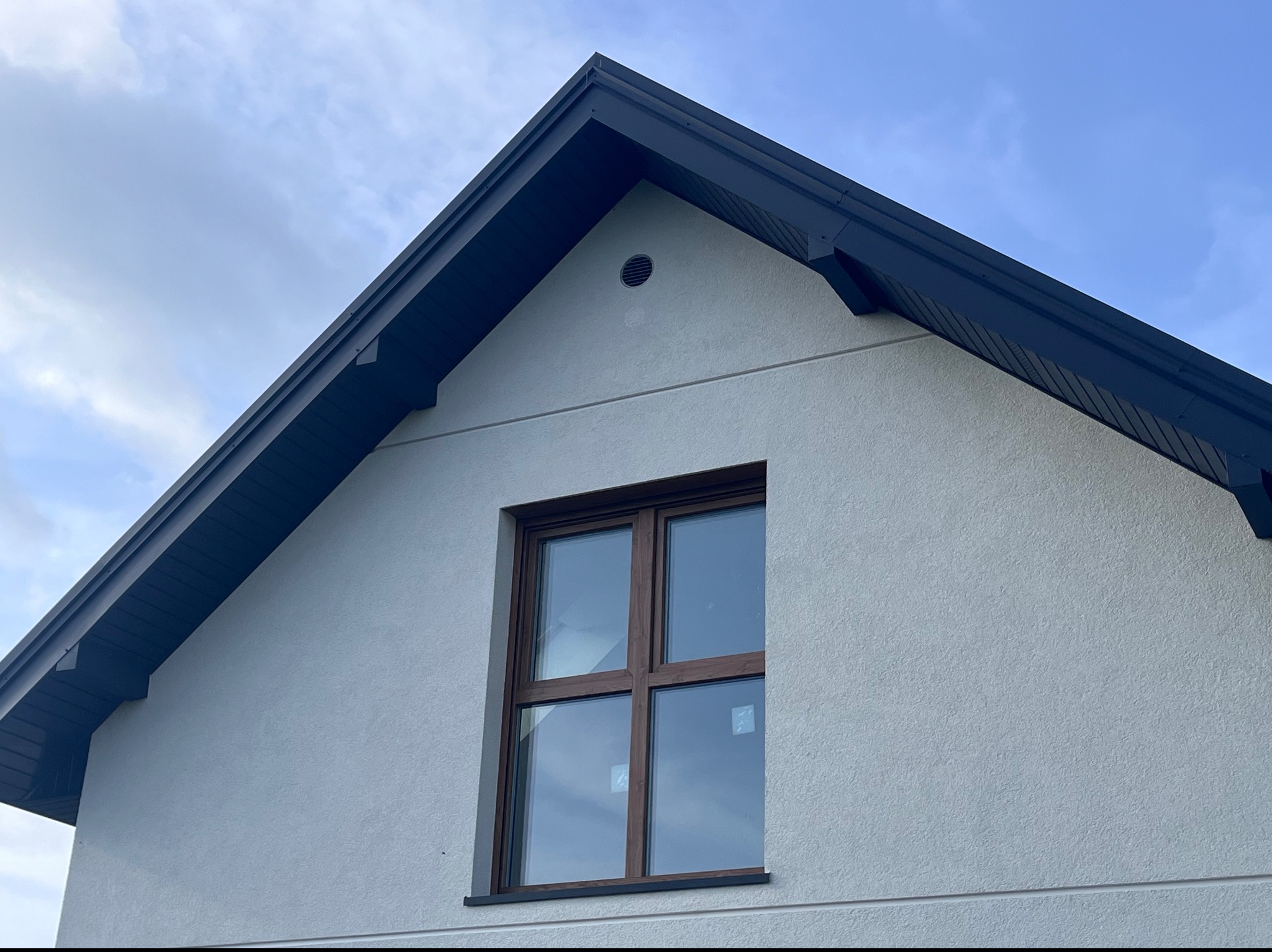 Fragment elewacji domu z ciemnym dachem i brązowym oknem, widoczny otwór wentylacyjny na szczycie ściany.