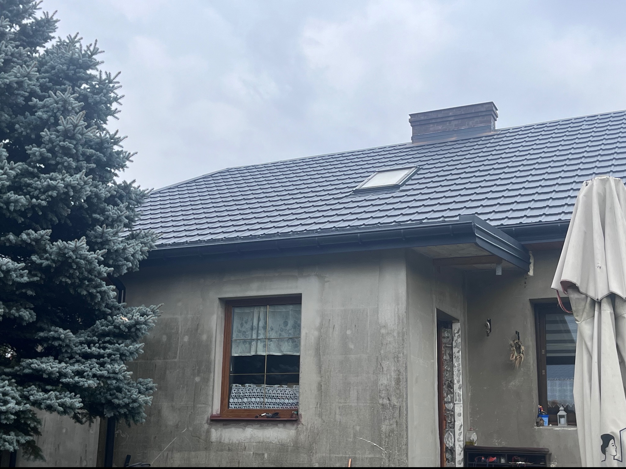 Dom z nowym, grafitowym dachem z dachówki, z oknem dachowym i kominem z cegły, widoczny fragment elewacji w trakcie prac wykończeniowych i srebrny świerk.