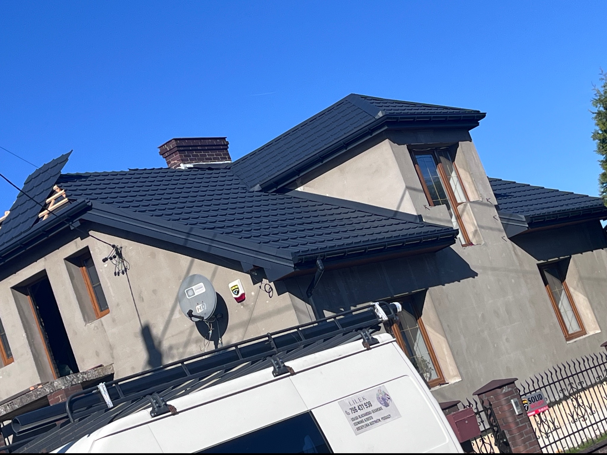 Dom z nowym, ciemnym dachem z dachówki bitumicznej, częściowo jeszcze w trakcie układania, widoczny komin i samochód firmy dekarskiej z reklamą.