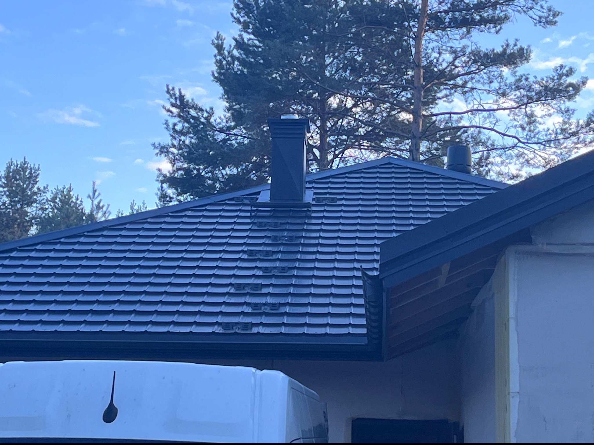 Nowy, ciemny dach z blachy trapezowej z kominem i wentylacją, widok z dołu, częściowo zasłonięty przez biały samochód dostawczy z czarnym logo firmy.