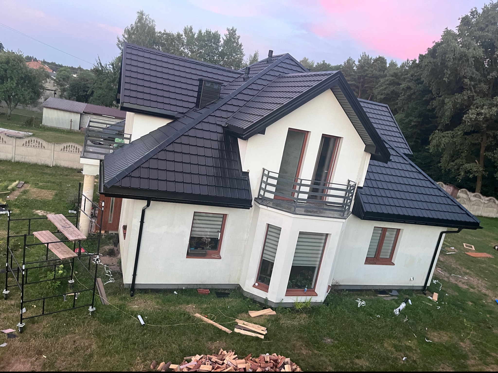 Nowoczesny dom jednorodzinny z nowym, czarnym dachem z blachodachówki, widoczne rusztowanie i przygotowane drewno opałowe na trawniku.