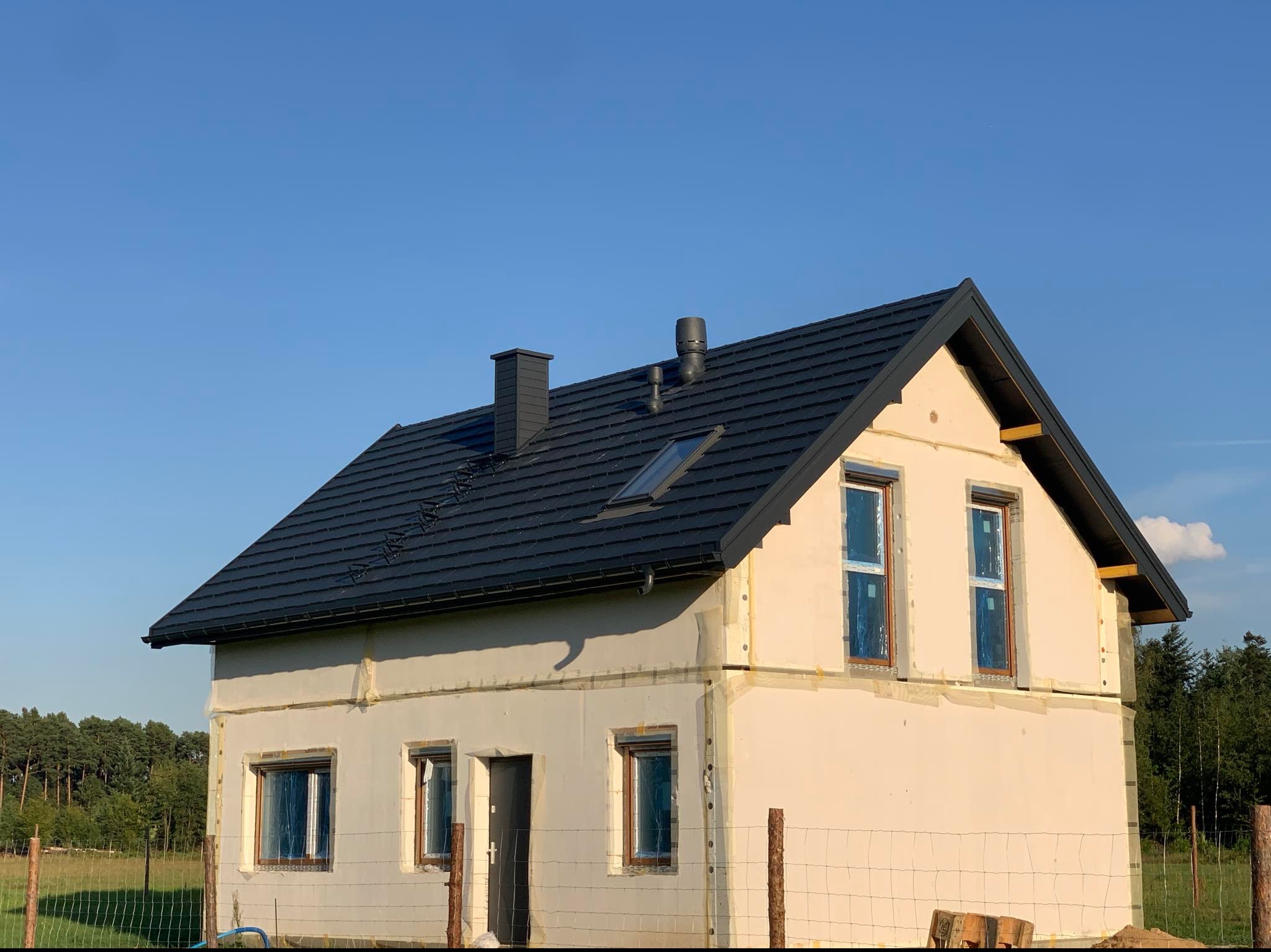 Dom jednorodzinny w trakcie ocieplania elewacji i układania ciemnej dachówki, widoczne kominy i okno dachowe, okna zabezpieczone folią, budynek otoczony siatką ogrodzeniową.