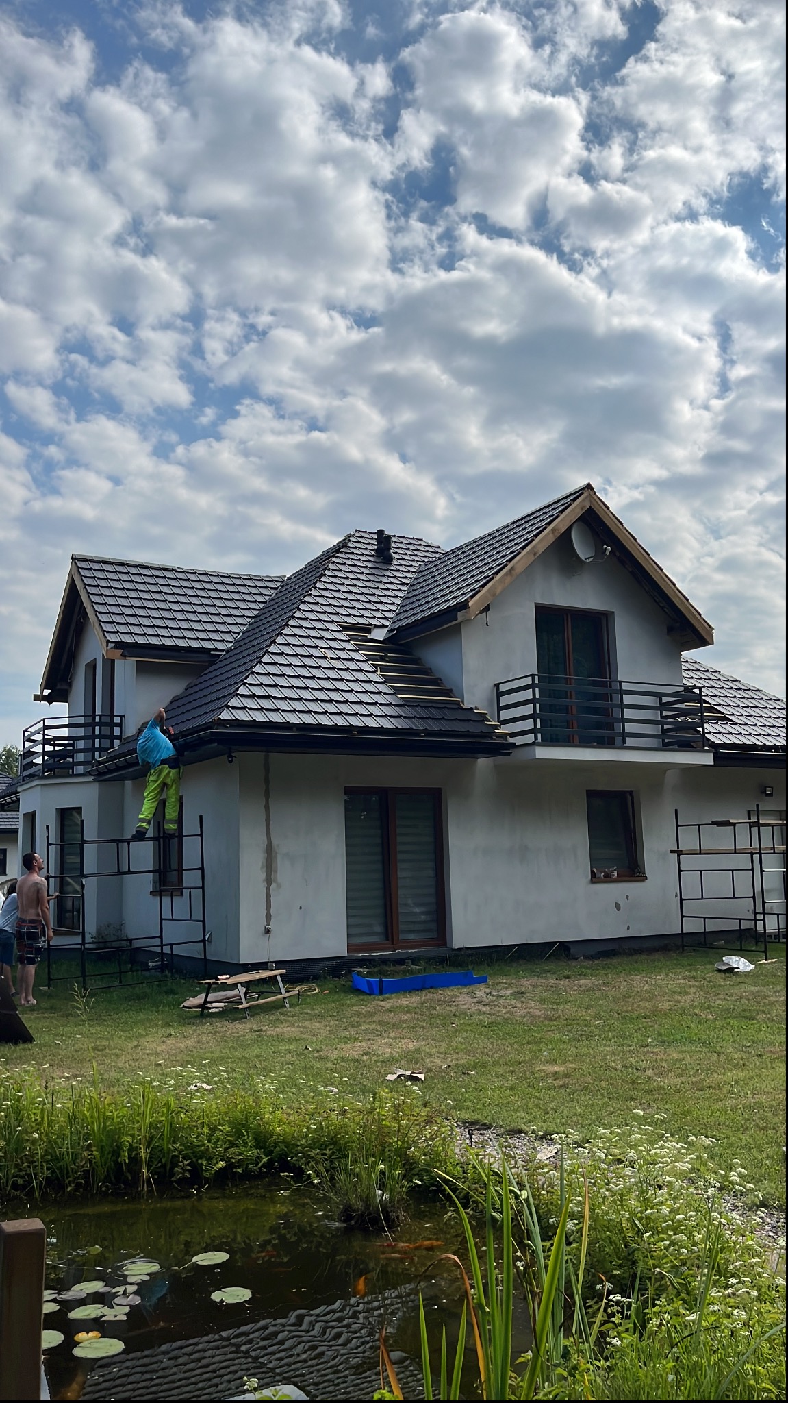 Dekarz w trakcie wymiany pokrycia dachu z czarnej dachówki na dwupiętrowym domu z balkonem, widoczne rusztowanie i staw z liliami wodnymi w ogrodzie.