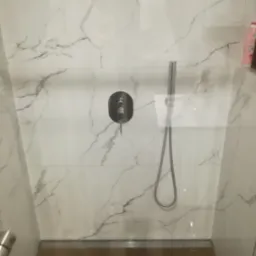 Szklana kabina prysznicowa z marmurowymi płytkami ściennymi, wyposażona w czarną baterię i srebrną słuchawkę prysznicową, z odpływem liniowym w podłodze wyłożonej panelami imitującymi drewno.