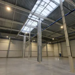 Wnętrze hali produkcyjnej z widocznymi instalacjami wentylacyjnymi pod sufitem, rurami w kolorze niebieskim oraz stalowymi słupami podtrzymującymi konstrukcję dachu z oknami.