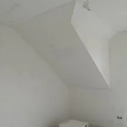 Wnętrze poddasza ze ścianami w trakcie szpachlowania, widoczne nierówności i niedoskonałości powierzchni przed ostatecznym wykończeniem, małe okno w prawym górnym rogu.