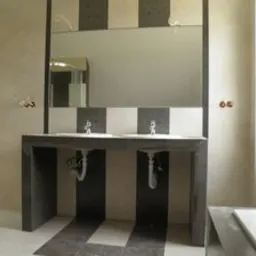 Łazienka w trakcie prac: podwójna umywalka na betonowym blacie, biało-szare płytki na ścianach i podłodze, widoczne rury pod umywalkami, duże lustro bez ramy.