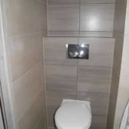 Zamontowana toaleta podwieszana z szarymi płytkami imitującymi drewno na ścianie i podłodze, widoczny chromowany przycisk spłuczki.