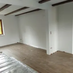 Układanie paneli podłogowych w pomieszczeniu z drewnianymi belkami stropowymi, widoczna folia izolacyjna i częściowo ułożone panele.