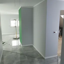 Korytarz w szarościach z wykończonymi podłogami z płytek imitujących kamień, widoczna lampa doświetlająca ścianę pomalowaną na zielono w sąsiednim pokoju, ściany przygotowane do malowania.