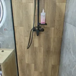 Kabina prysznicowa z deszczownicą w kolorze czarnym, wyłożona płytkami imitującymi drewno i szarym marmurem, z odpływem liniowym i półką z kosmetykiem.