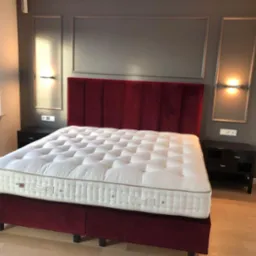 Sypialnia z ł&oacute;żkiem z bordowym, welurowym zagł&oacute;wkiem i ramą, białym materacem z pikowaniem, drewnianą podłogą i szarymi ścianami z dekoracyjnymi listwami i kinkietami.