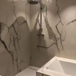 Nowoczesna łazienka z kabiną prysznicową typu walk-in i wanną obudowaną płytami imitującymi marmur, oświetlenie sufitowe halogenowe.