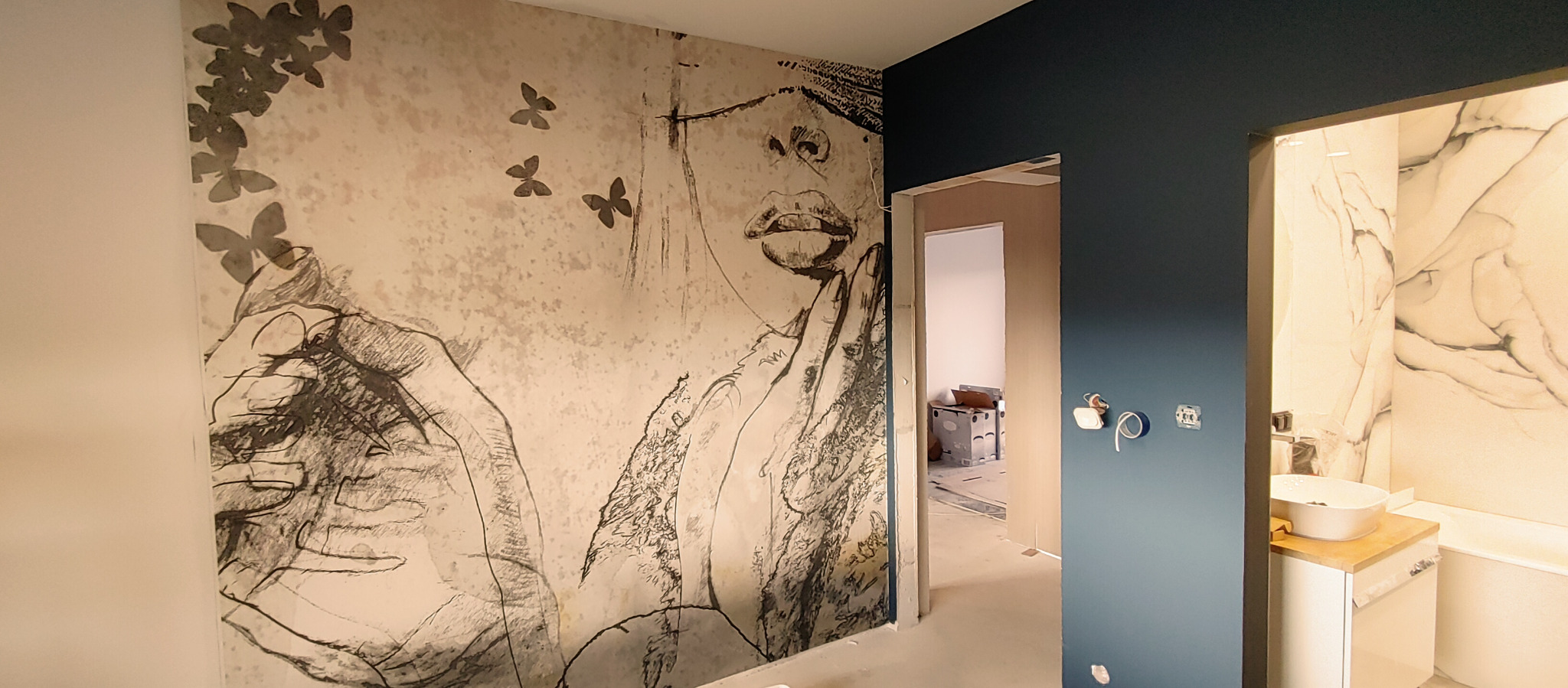 Ściana z motywem kobiety i motyli w pokoju, widok na wykończoną łazienkę z białą umywalką nablatową i płytkami imitującymi marmur.