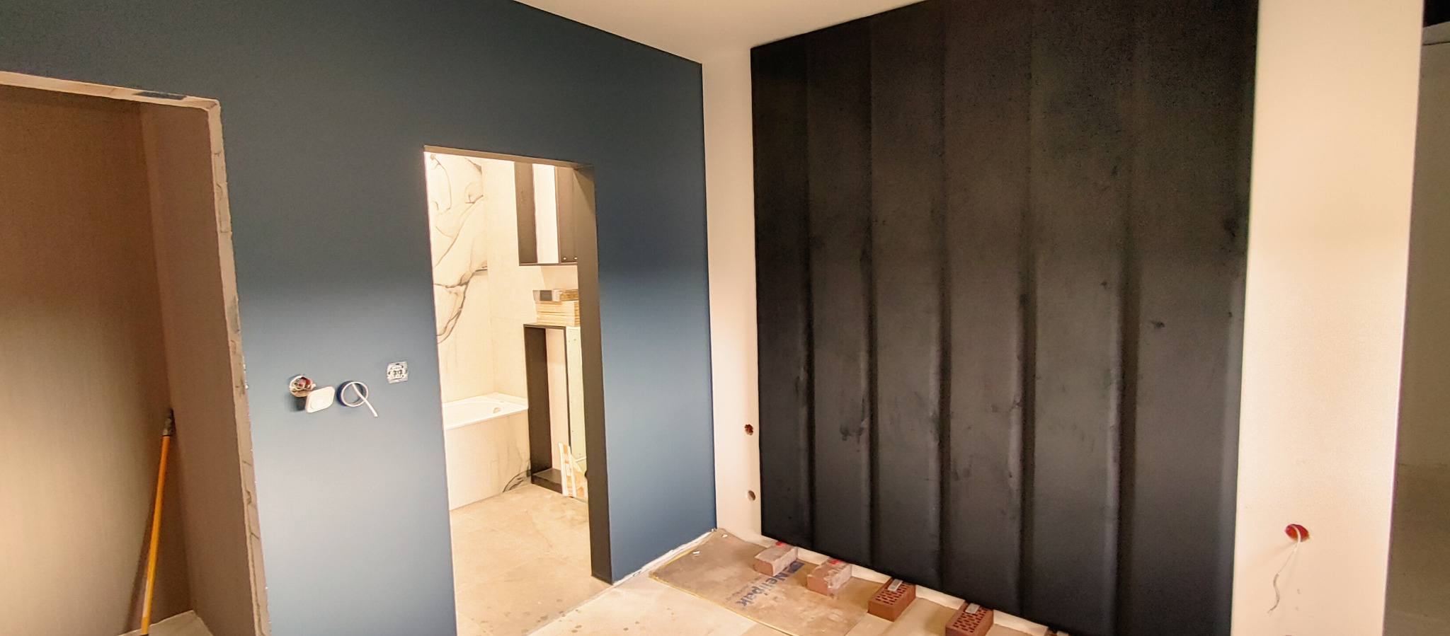 Niedokończony remont łazienki: widok na wannę z marmurowymi płytkami przez otwór w ścianie, niebieska ściana z gniazdkami, czarna ściana z pionowymi panelami i cegły na podłodze.