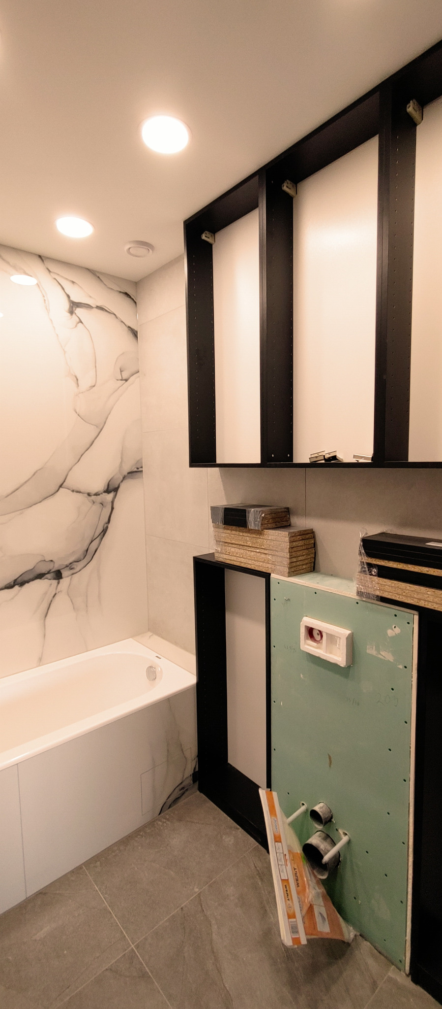 Łazienka w trakcie montażu: biała wanna z marmurowym wzorem na ścianie, czarne ramy szafek bez frontów, płyty kartonowo-gipsowe z instalacją hydrauliczną, stosy płyt meblowych na szafkach.