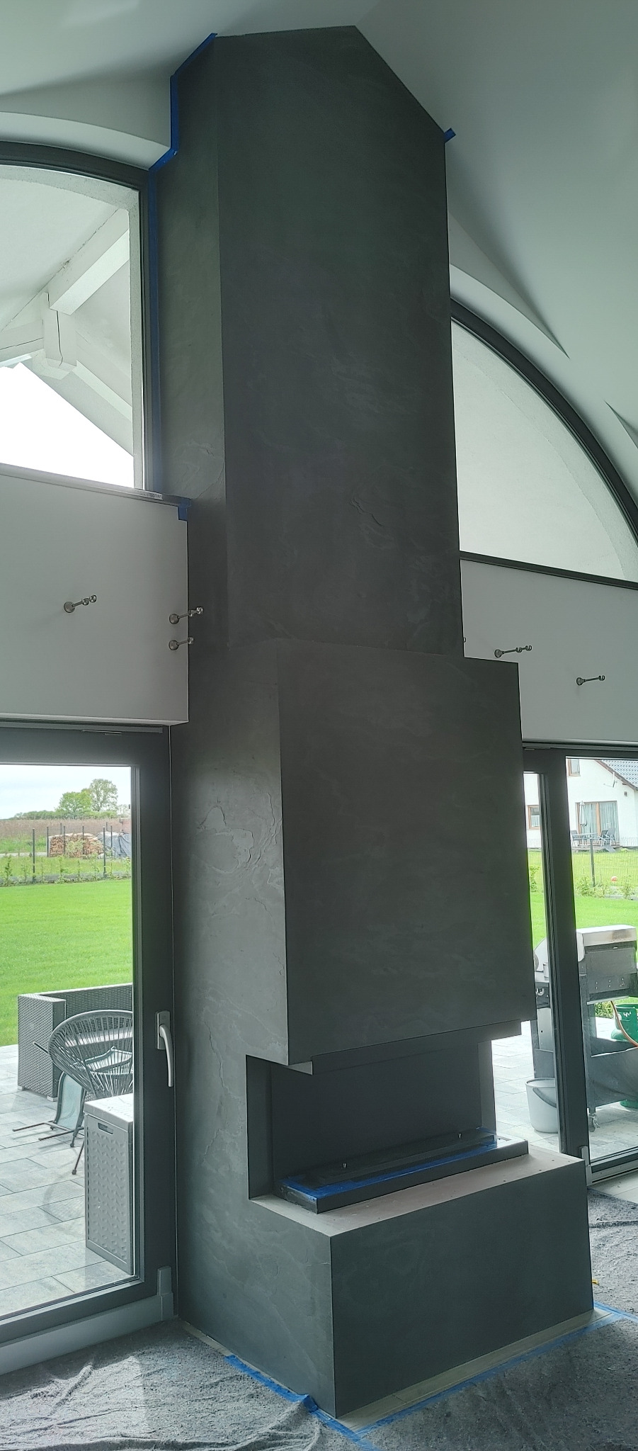 Wysoki komin z dekoracyjną szarą strukturą, zabezpieczony taśmą malarską podczas prac wykończeniowych, widoczny z perspektywy podłogi w jasnym pomieszczeniu z dużymi oknami i widokiem na ogród.