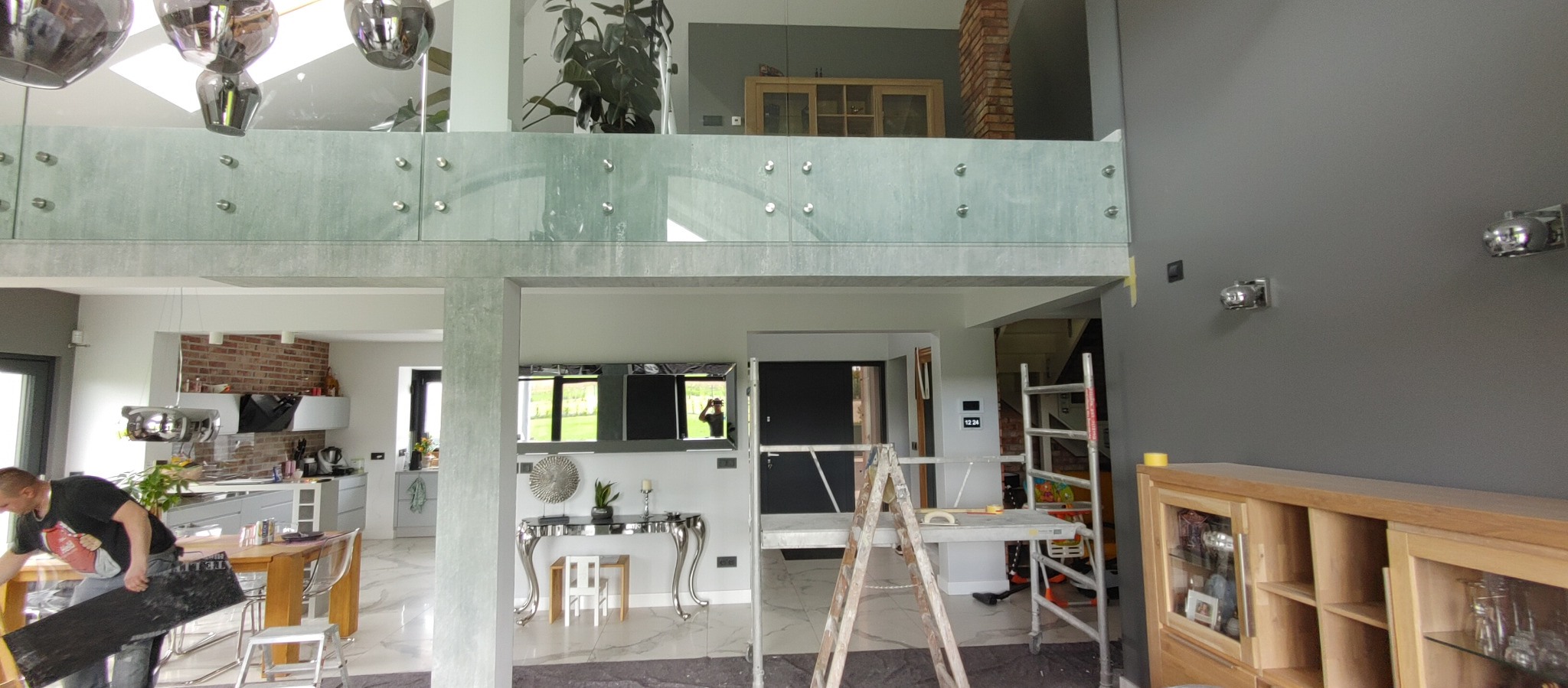 Dwupoziomowe wnętrze domu w trakcie odświeżania: widoczny fragment kuchni z ceglaną ścianą, salon z konsolą w stylu glamour i drabina malarska przy szarej ścianie z zamontowanymi lampami.