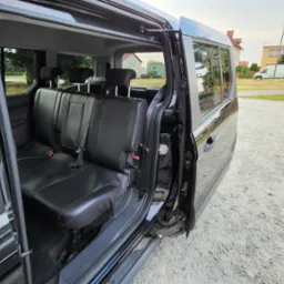 Wnętrze czarnego vana z otwartymi drzwiami przesuwnymi, ukazujące czarne skórzane siedzenia i widok na budynki mieszkalne w tle.