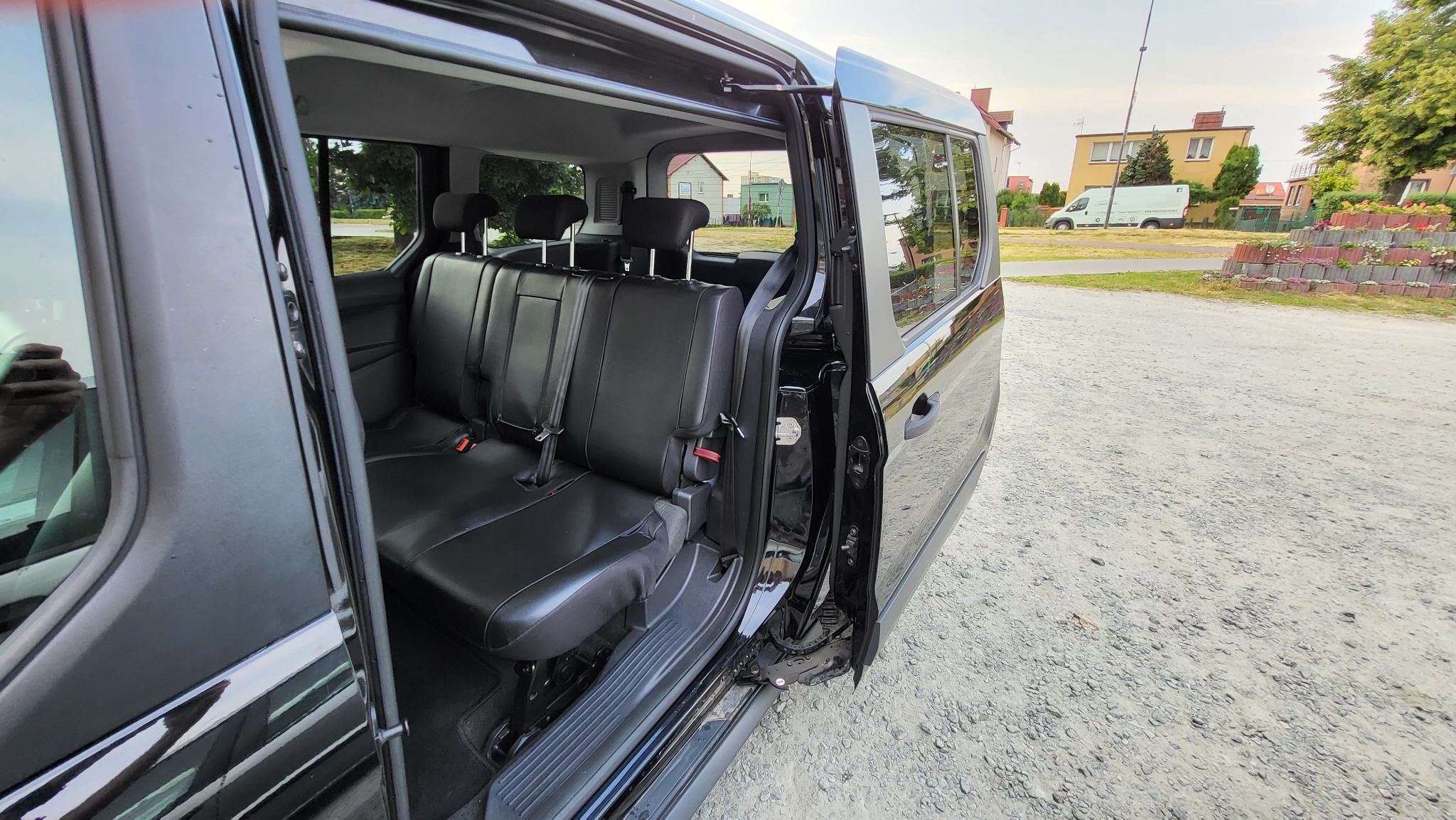 Wnętrze czarnego vana z otwartymi drzwiami przesuwnymi, ukazujące czarne skórzane siedzenia i widok na budynki mieszkalne w tle.