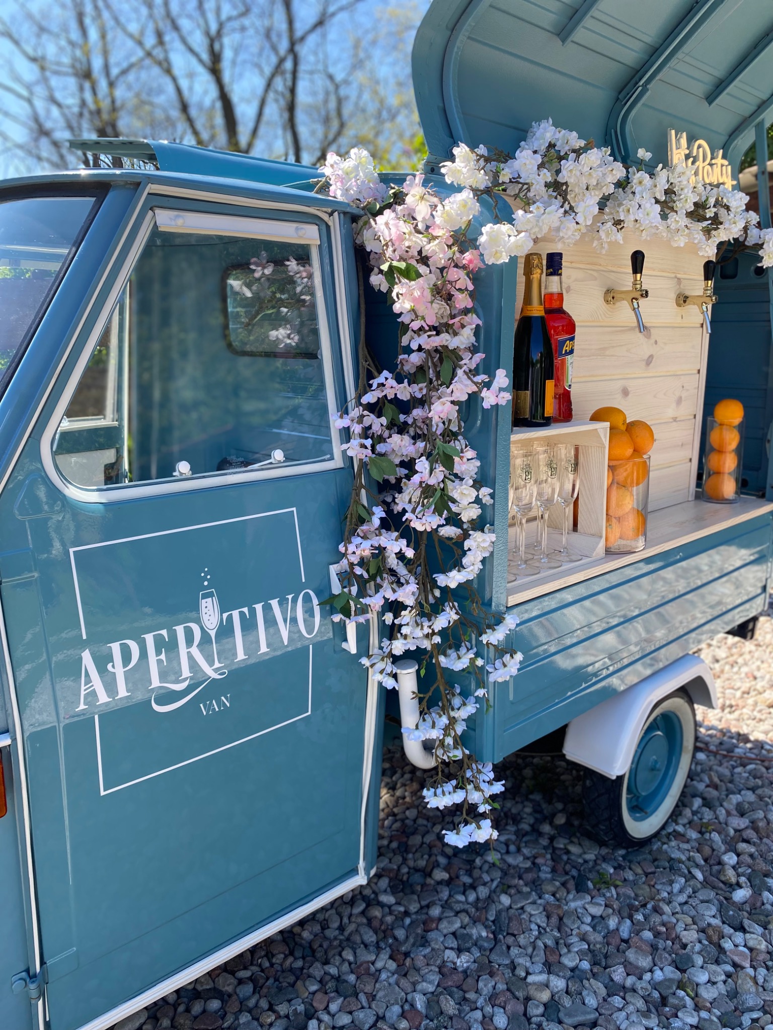 Niebieski, mobilny bar 'Aperitivo Van' udekorowany białymi i różowymi kwiatami, z butelkami alkoholu, szklankami i pomarańczami, gotowy do serwowania drinków na imprezie.