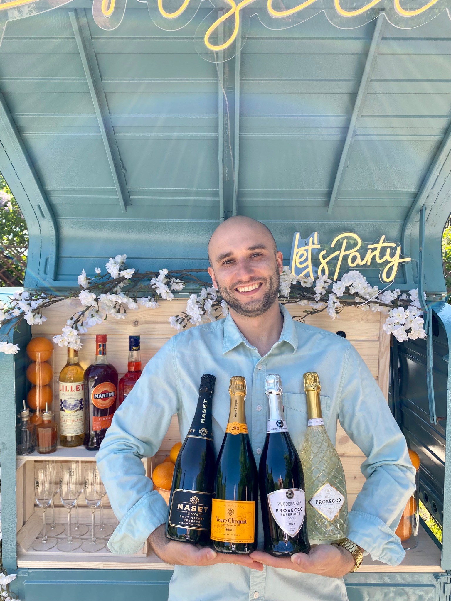 Uśmiechnięty barman prezentuje cztery butelki szampana i prosecco przed mobilnym barem z napisem 'Let's Party', udekorowanym pomarańczami i białymi kwiatami.