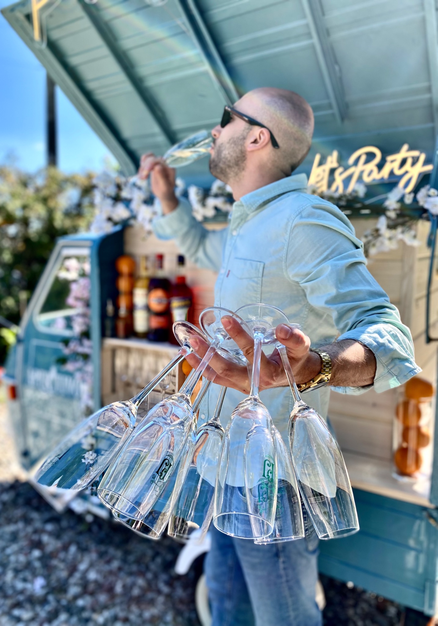 Mężczyzna w okularach pije szampana, trzymając w dłoniach kilka pustych kieliszków do szampana przed mobilnym barem z napisem 'Let's Party' i kwiatową dekoracją.