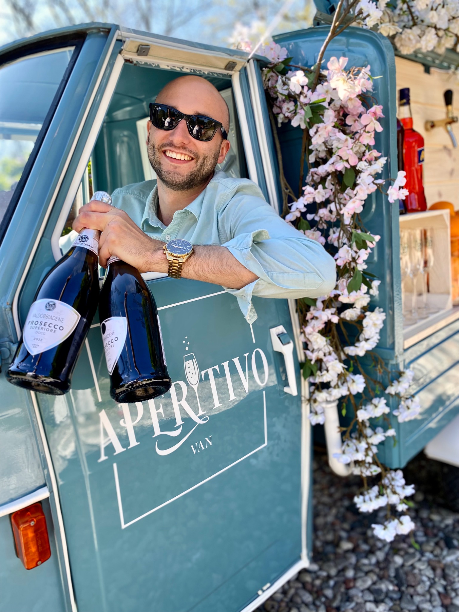 Uśmiechnięty mężczyzna w okularach słonecznych, wystający z niebieskiego wozu z napisem 'Aperitivo Van', trzymający dwie butelki Prosecco Superiore, z dekoracją z kwiatów wiśni.