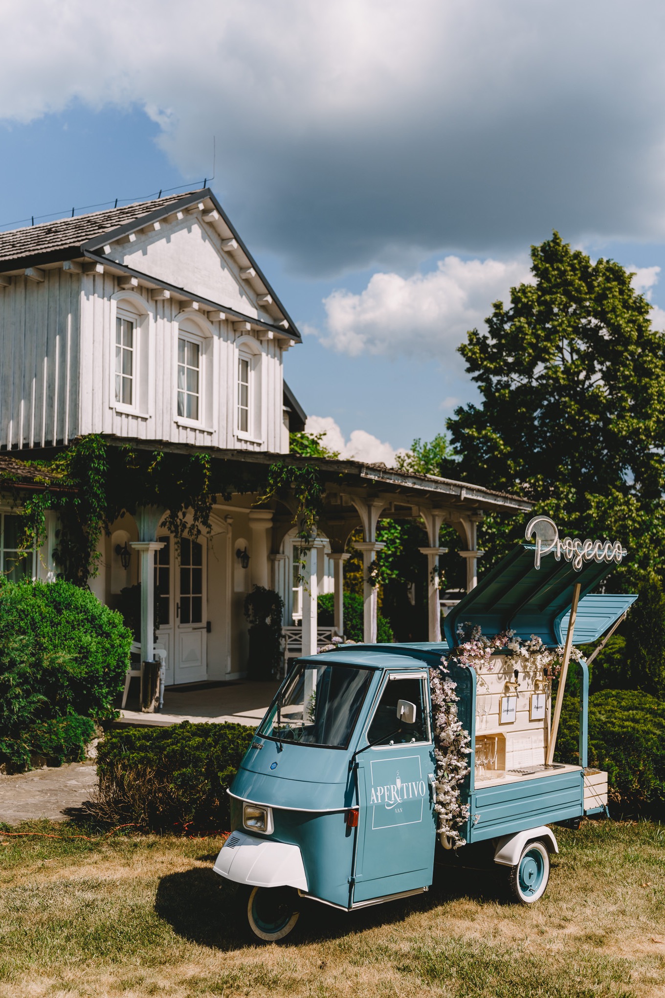 Niebieski trójkołowy bar mobilny 'Aperitivo Van' z napisem 'Prosecco' na tle białego domu z kolumnami, udekorowany kwiatami.