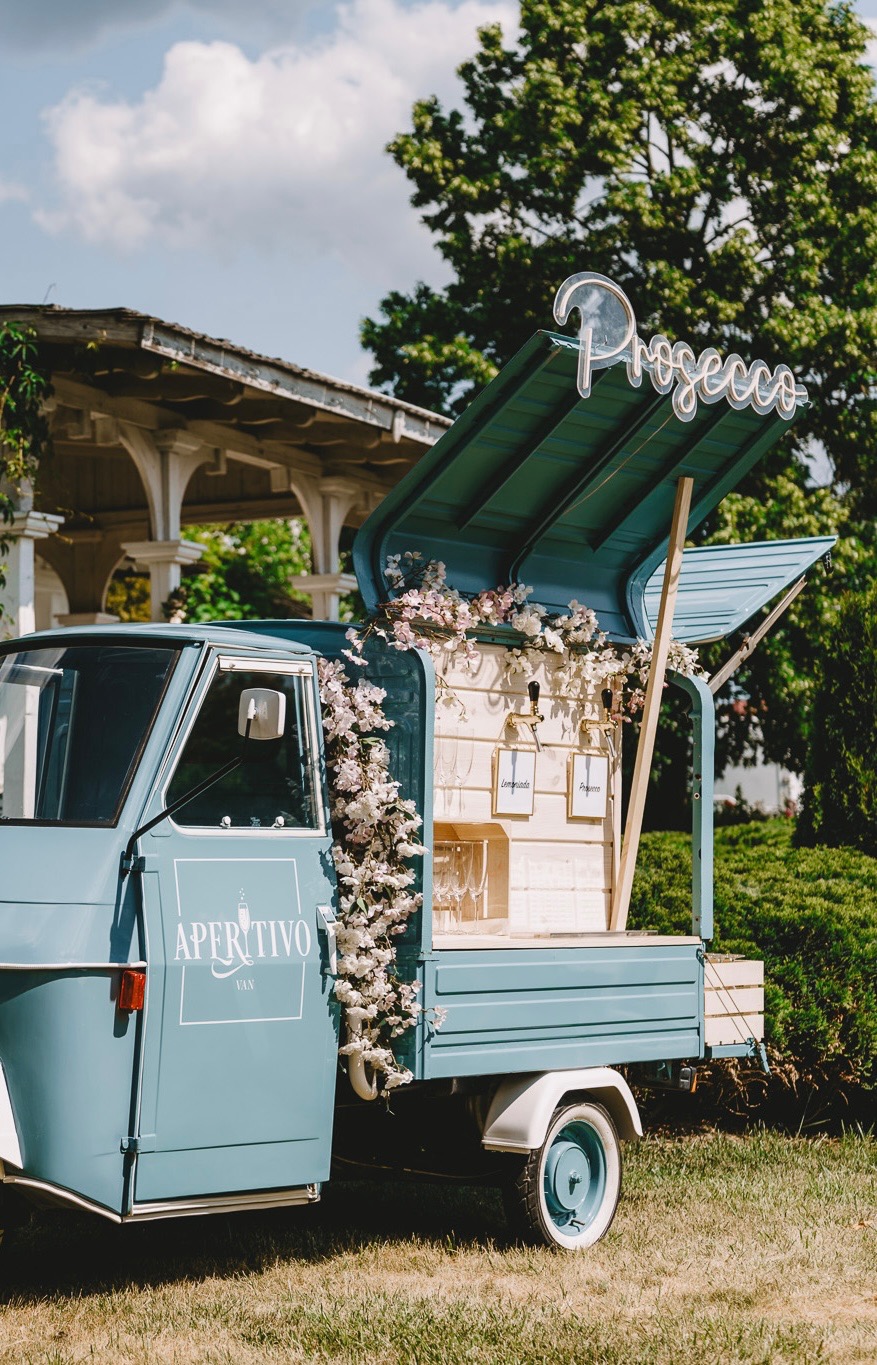 Niebieski Ape Van z napisem 'Aperitivo Van', przerobiony na mobilny bar z prosecco i lemoniadą, ozdobiony kwiatami, na tle zieleni.
