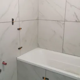 Układanie białych płytek imitujących marmur w łazience, widoczne pomarańczowe krzyżyki dystansowe, częściowo wykończona obudowa wanny.