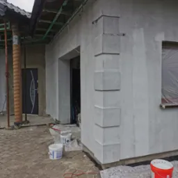 Świeżo otynkowana elewacja domu jednorodzinnego z dekoracyjnymi narożnikami, widoczne wiaderka z farbą i narzędzia malarskie, okno zabezpieczone folią malarską.