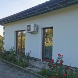 Świeżo pomalowana, jasna elewacja domu jednorodzinnego z widocznym klimatyzatorem marki Heiko, brązowe ramy okienne, ciemna dachówka i rabata z czerwonymi różami wzdłuż ściany.