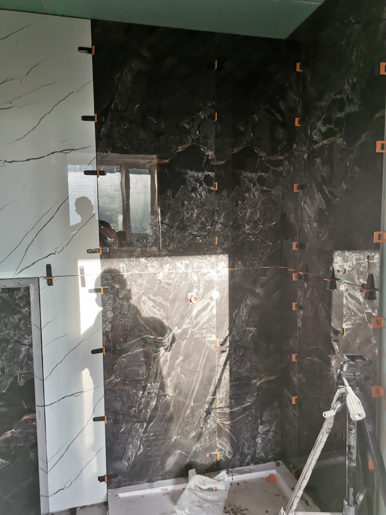 Układanie płytek imitujących marmur w łazience w Chełmie Śląskim, widoczne białe i czarne płytki z użyciem klipsów poziomujących, cień osoby w odbiciu płytek.