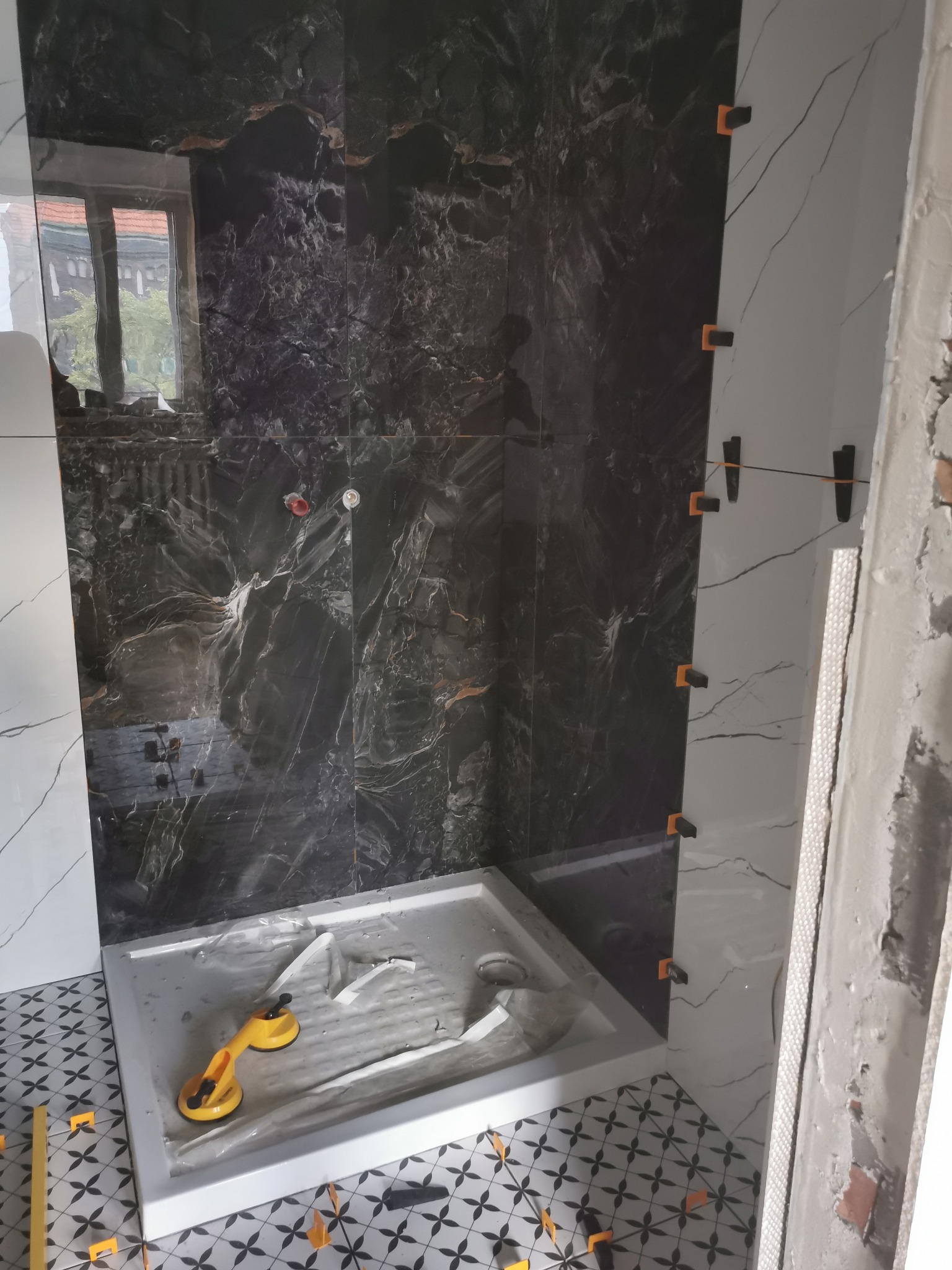 Montaż czarno-białych płytek imitujących marmur w kabinie prysznicowej, widoczne przyssawki do płytek i poziomowanie krzyżykami.