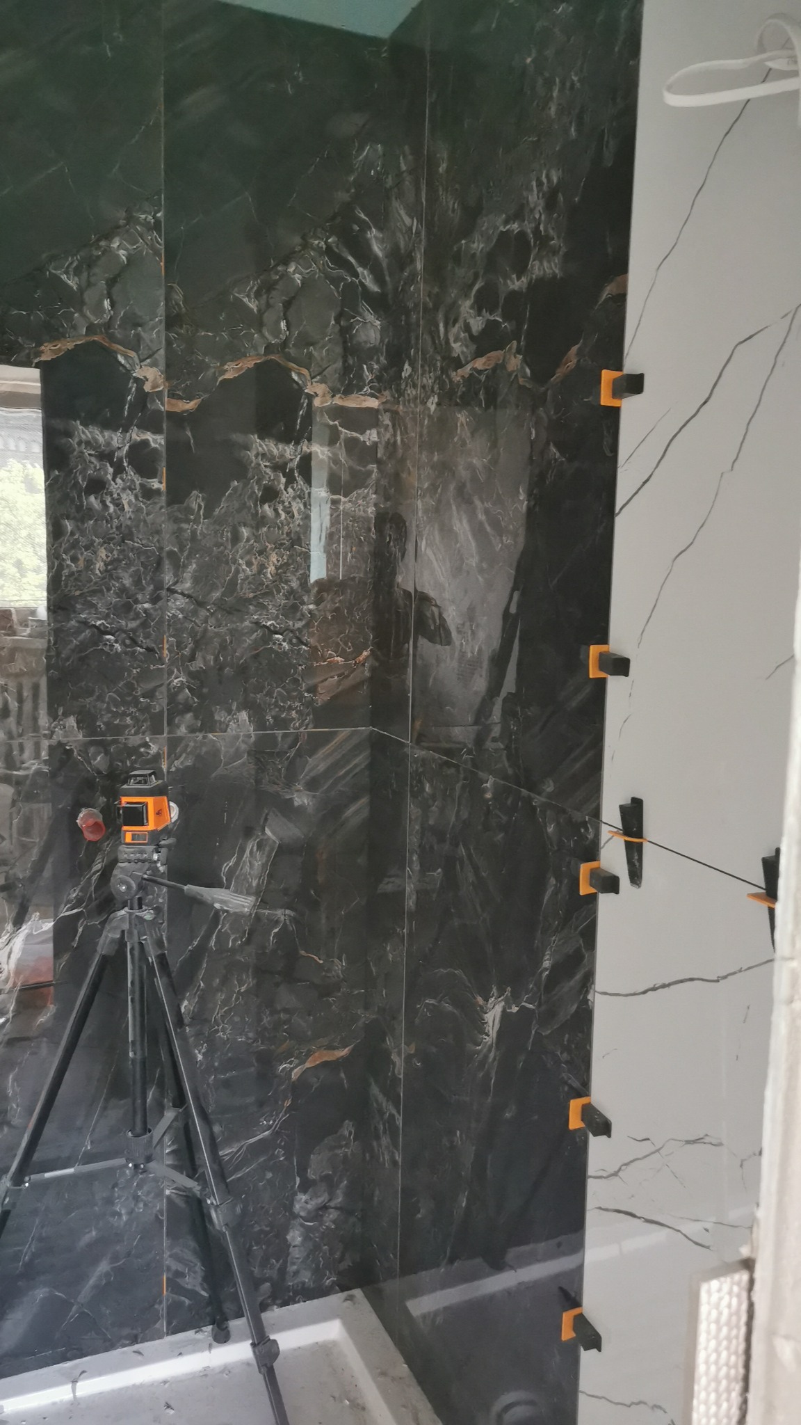 Układanie płytek imitujących czarny marmur na ścianie łazienki, widoczny laser krzyżowy na statywie oraz czarne kliny dystansowe na białych płytkach.