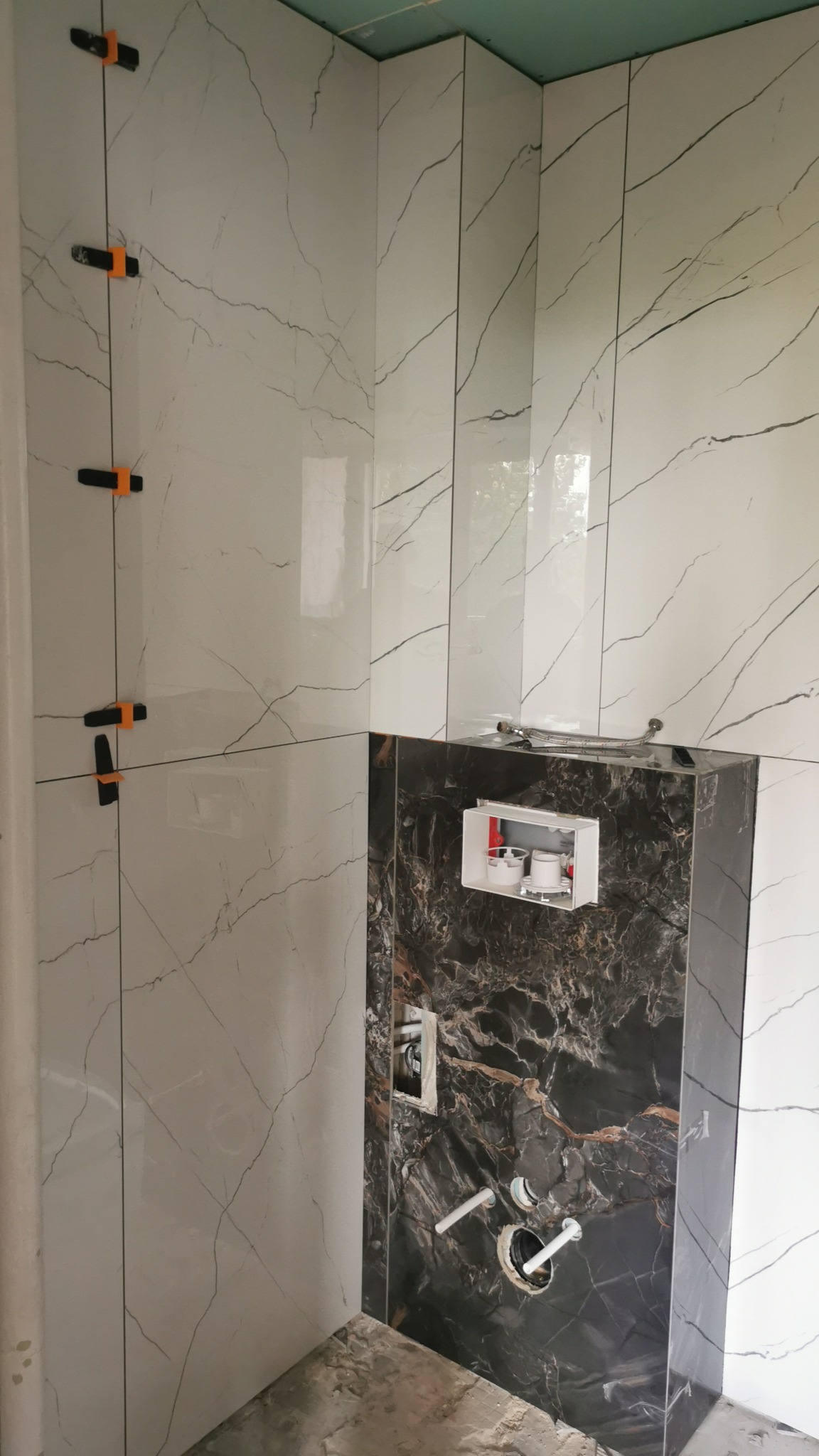 Ściana łazienki w trakcie wykańczania, z białymi płytkami imitującymi marmur, czarne płytki z widoczną instalacją podtynkową WC, pomarańczowe klipsy poziomujące płytki.