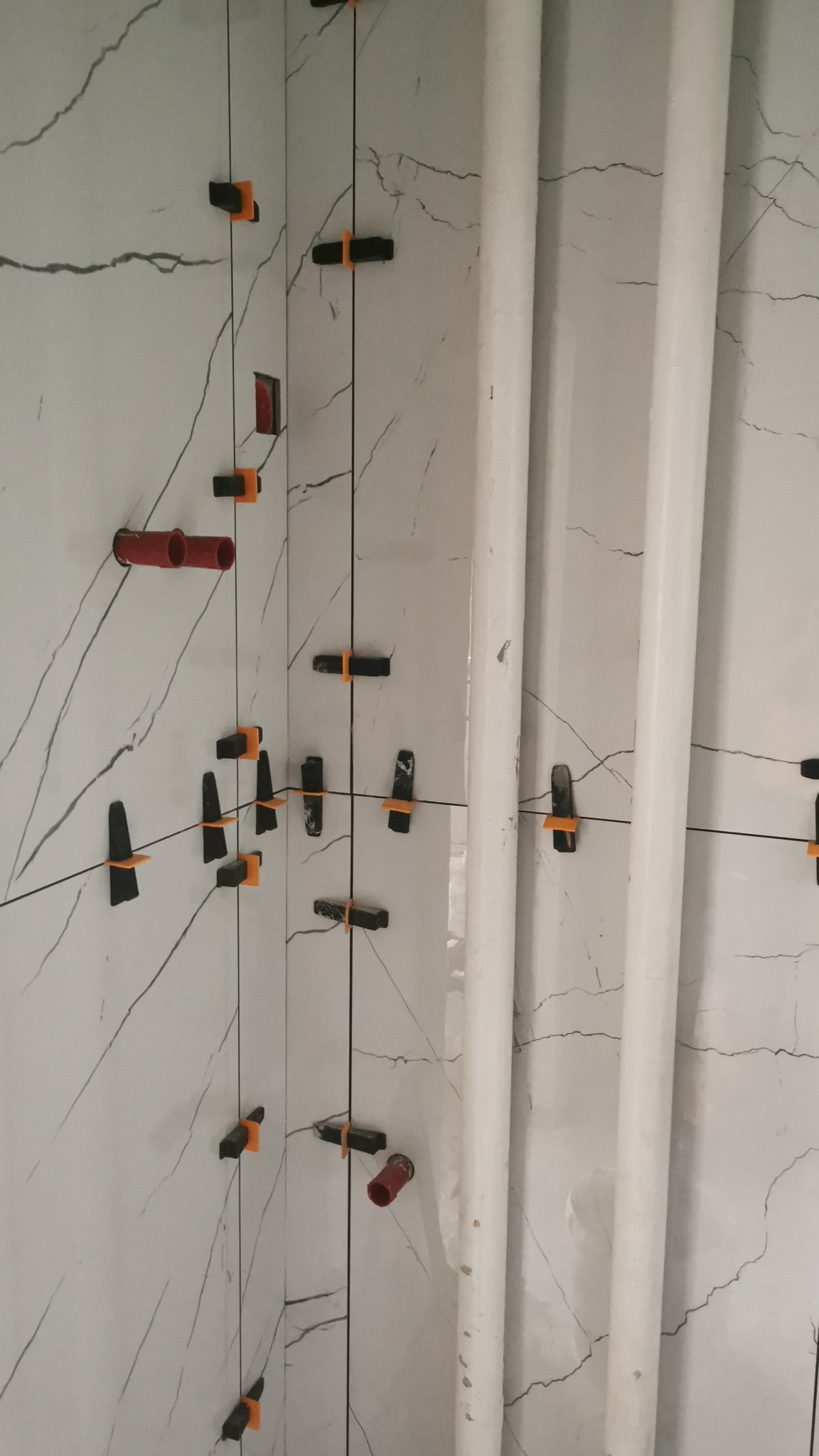 Ściana w trakcie układania płytek imitujących marmur, z widocznymi rurami i systemem poziomowania płytek z czarnymi klipsami i pomarańczowymi klinami.