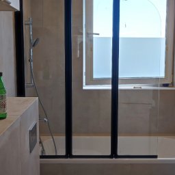 Kompleksowe wykończenie wnętrz Serhii Bakharev - Wykończona łazienka z wanną i szklanym parawanem z czarnymi profilami. Na pierwszym planie toaleta zabezpieczona folią ochronną. Widoczne beżowe płytki i marmurowa podłoga.