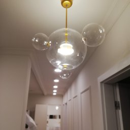 Kompleksowe wykończenie wnętrz Serhii Bakharev - Szklany żyrandol w kształcie kuli z trzema mniejszymi kulami, zawieszony na złotym pręcie w białym korytarzu z widokiem na łazienkę w tle.