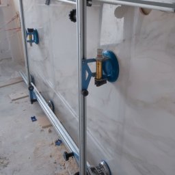 Kompleksowe wykończenie wnętrz Serhii Bakharev - Montaż wielkoformatowej płyty imitującej marmur za pomocą specjalistycznego systemu szyn i przyssawek, widoczne narzędzia i drobne zabrudzenia na podłodze.