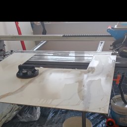 Kompleksowe wykończenie wnętrz Serhii Bakharev - Płytka ceramiczna z marmurkowym wzorem, przycięta z otworem, leży na stole roboczym z przyssawką, obok stoi profesjonalna przecinarka do płytek, na podłodze folia ochronna.