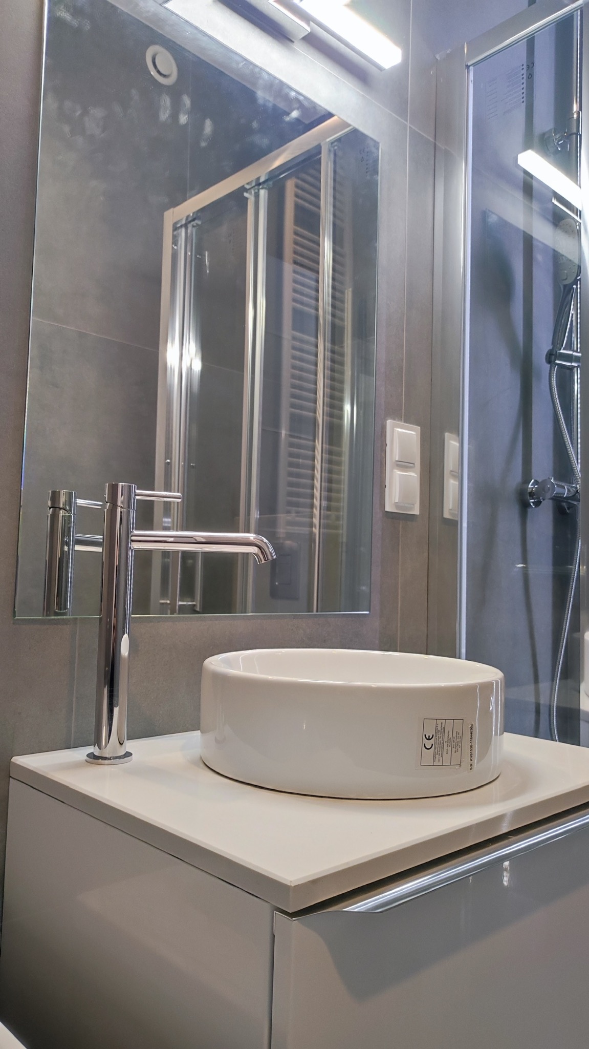 Nowoczesna łazienka: biała, okrągła umywalka nablatowa z chromowaną baterią na szafce, lustro z odbiciem kabiny prysznicowej, szare płytki.