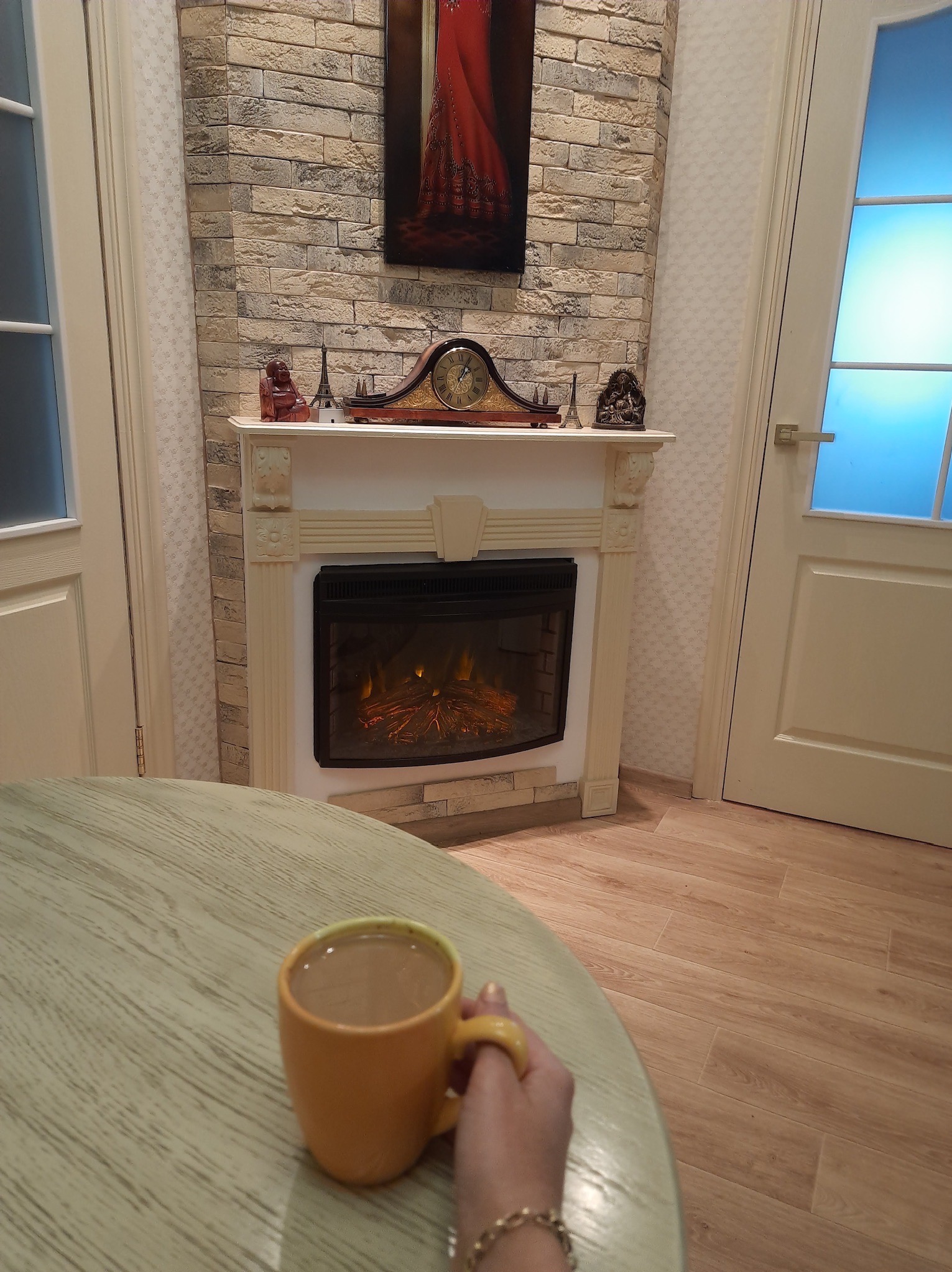 Elegancki salon z elektrycznym kominkiem obudowanym dekoracyjną cegłą, ozdobami i obrazem. Na pierwszym planie filiżanka kawy na okrągłym stole.