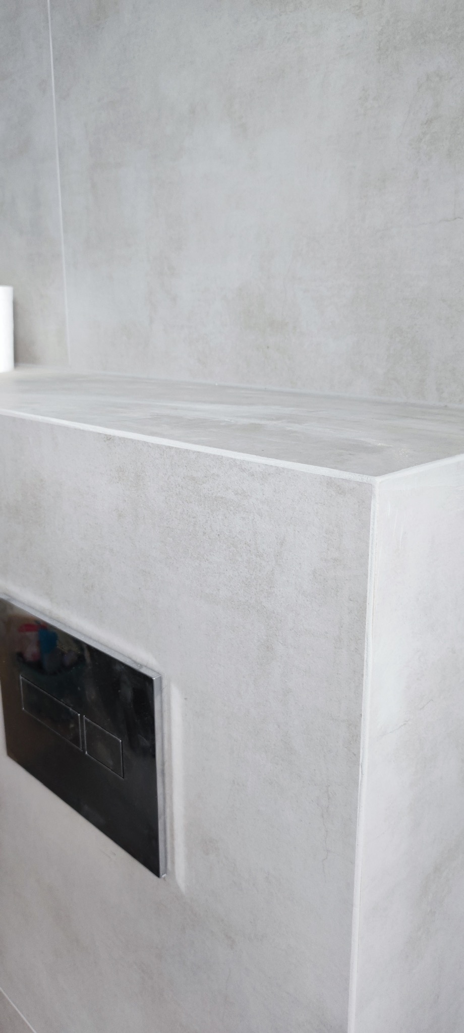 Minimalistyczna łazienka: szara, matowa płytka imitująca beton, zabudowa WC z chromowanym przyciskiem. Estetyczne wykończenie narożników.