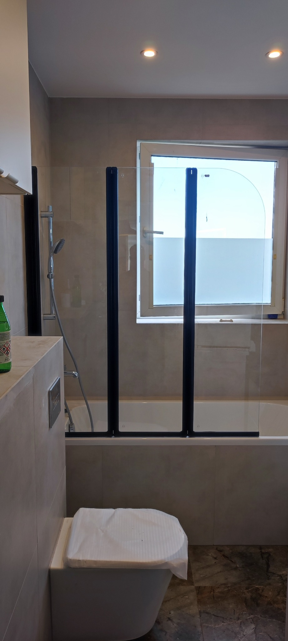 Wykończona łazienka z wanną i szklanym parawanem z czarnymi profilami. Na pierwszym planie toaleta zabezpieczona folią ochronną. Widoczne beżowe płytki i marmurowa podłoga.