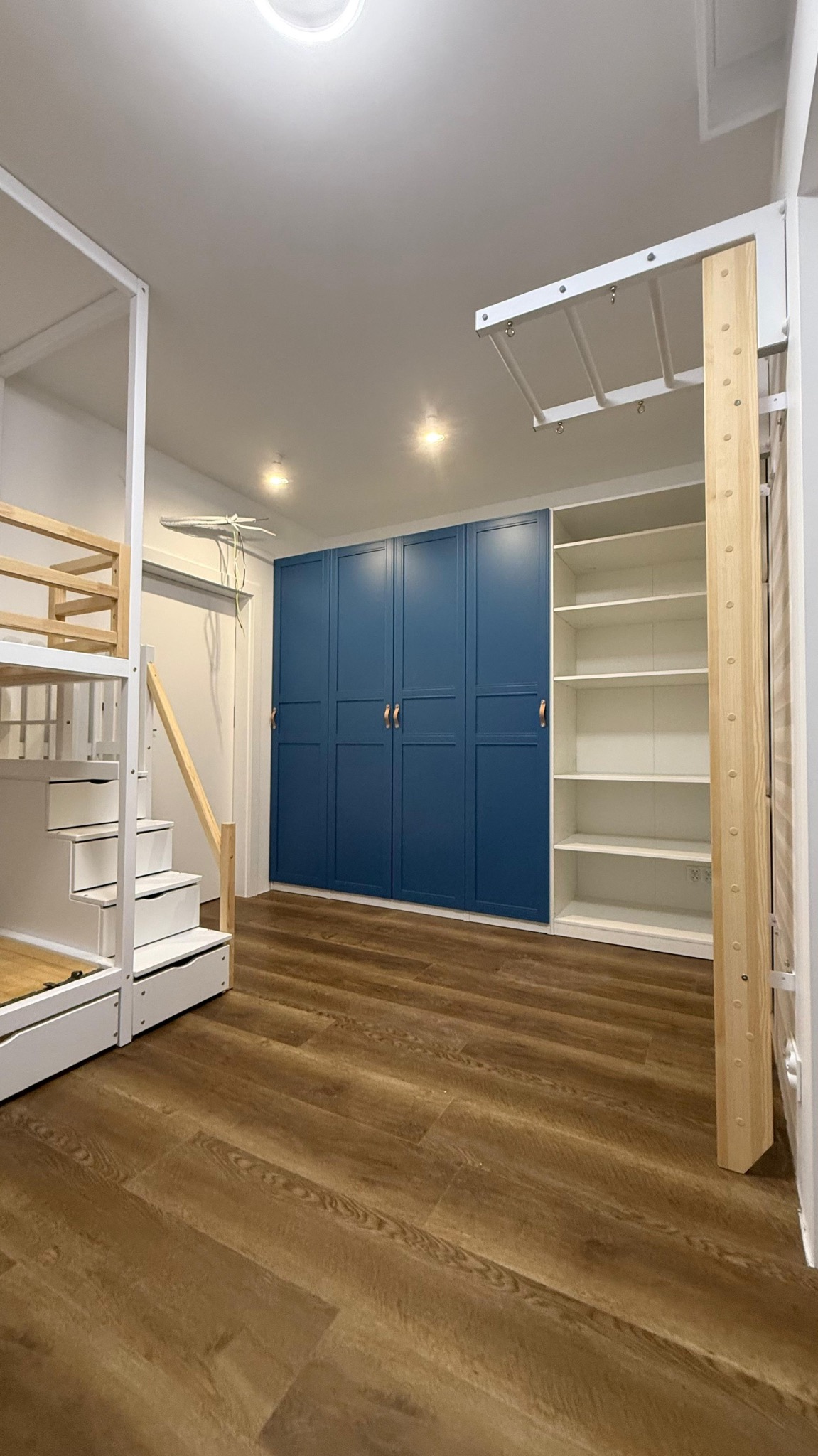 Pokój dziecięcy po remoncie: drewniana podłoga, zabudowa z szafami w kolorze granatowym, białe regały i łóżko piętrowe z drabinką.