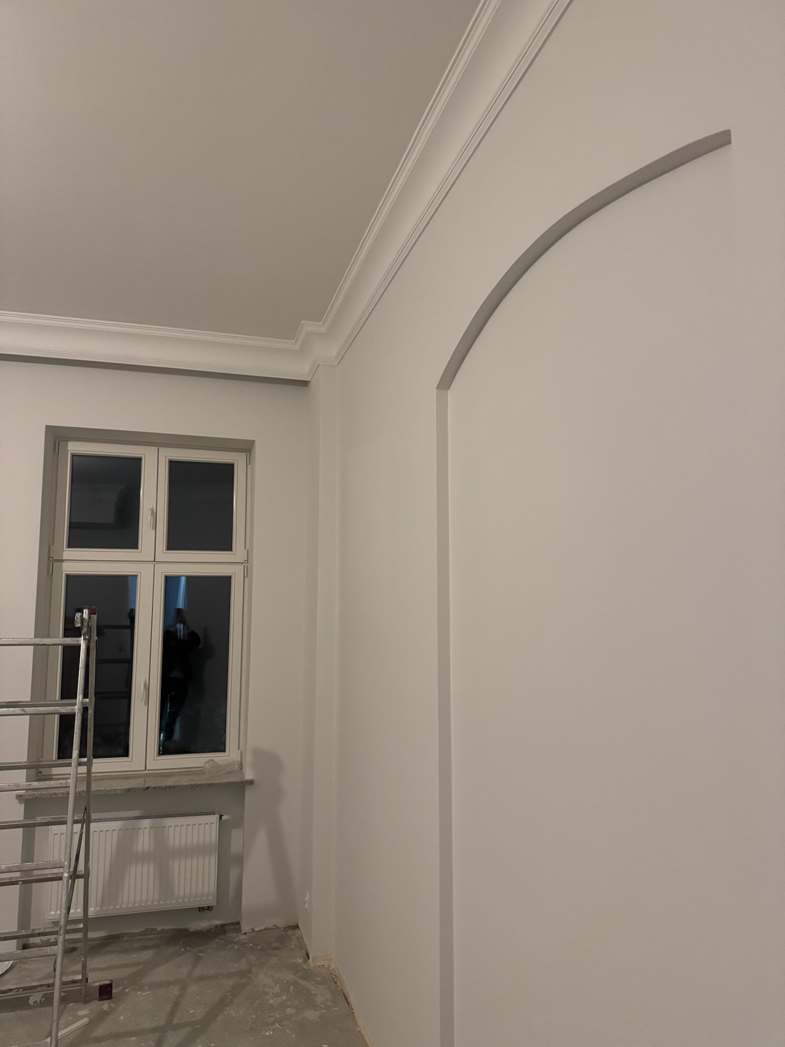 Białe wnętrze po remoncie: gładkie ściany z łukowatym przejściem, okno z parapetem i grzejnikiem. Aluminiowa drabina oparta o ścianę. Listwy dekoracyjne pod sufitem.
